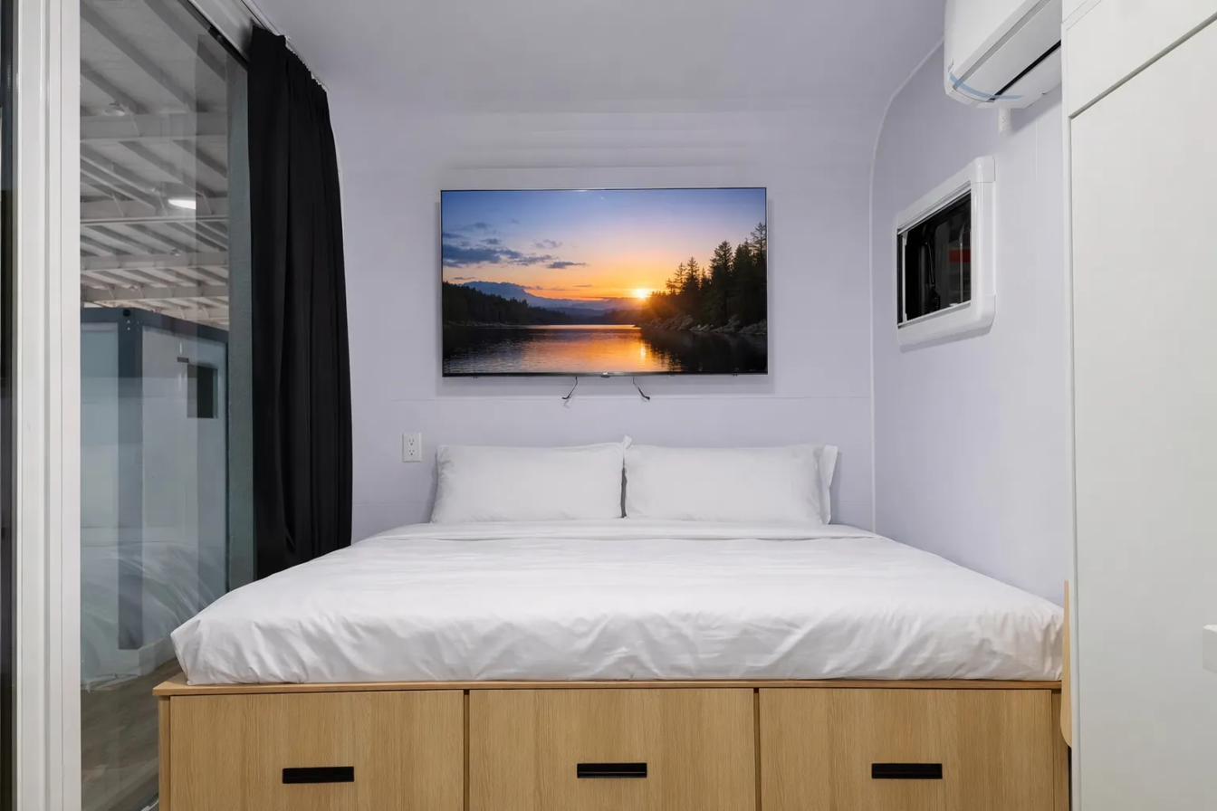 Standard 20ft tiny house bedroom — queen bed with storage drawers and wall-mounted TV showing sunset over lake
